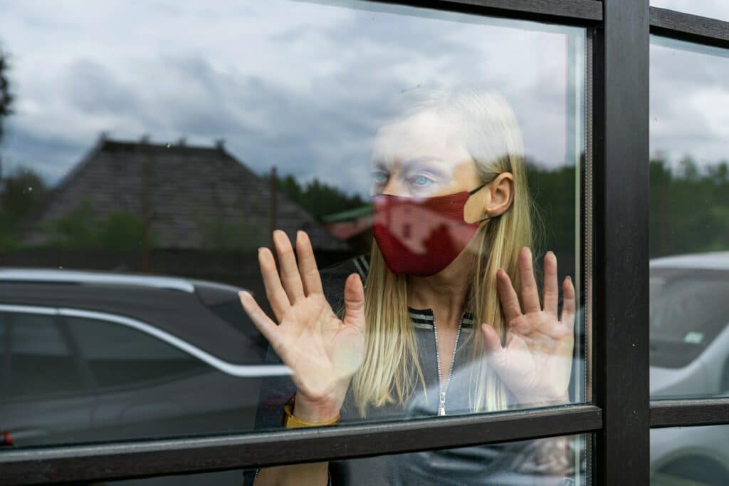 depresive woman in face mask looking out the window, staying home in quarantine, covid-19 lockdown